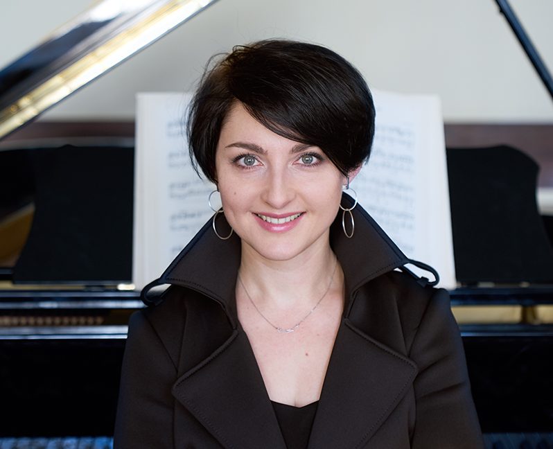Photo of Agnieszka Lasko sitting at a grand piano with her back to the keyboard and smiling at the camera.